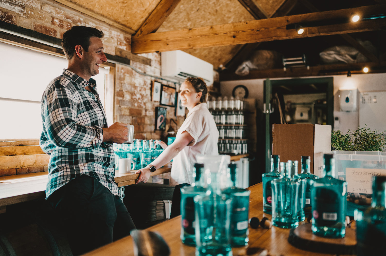 Emily and Rupert share a glance in the distillery with a cup of tea. Bottles on the table and stacked up in production