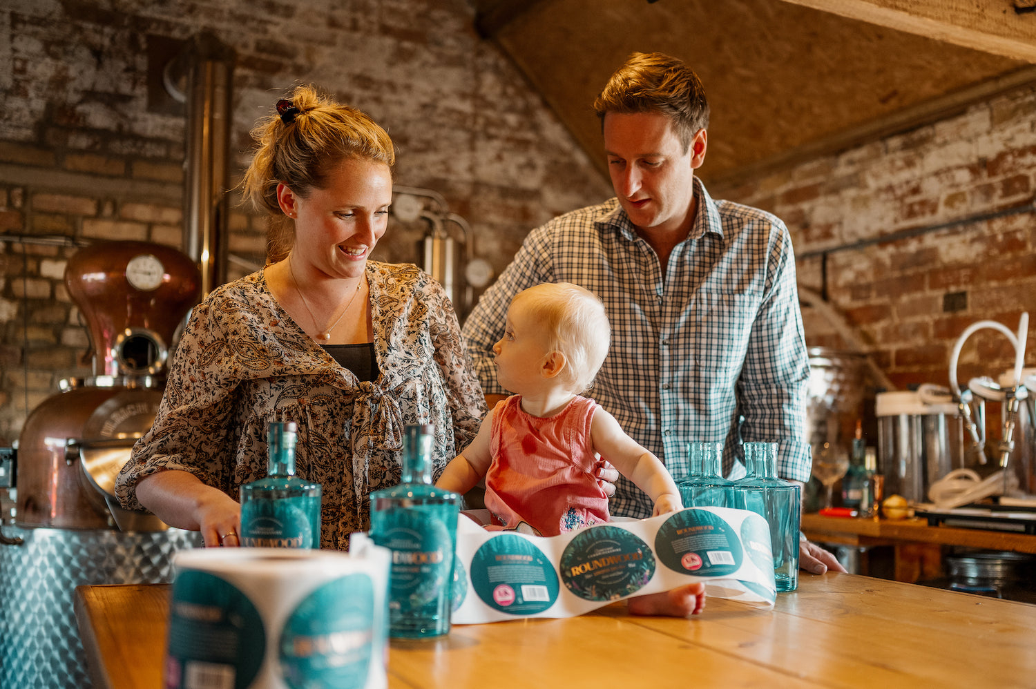 Roundwood Family - Emily and Rupert laugh with baby daughter perched on table surrounded by labels