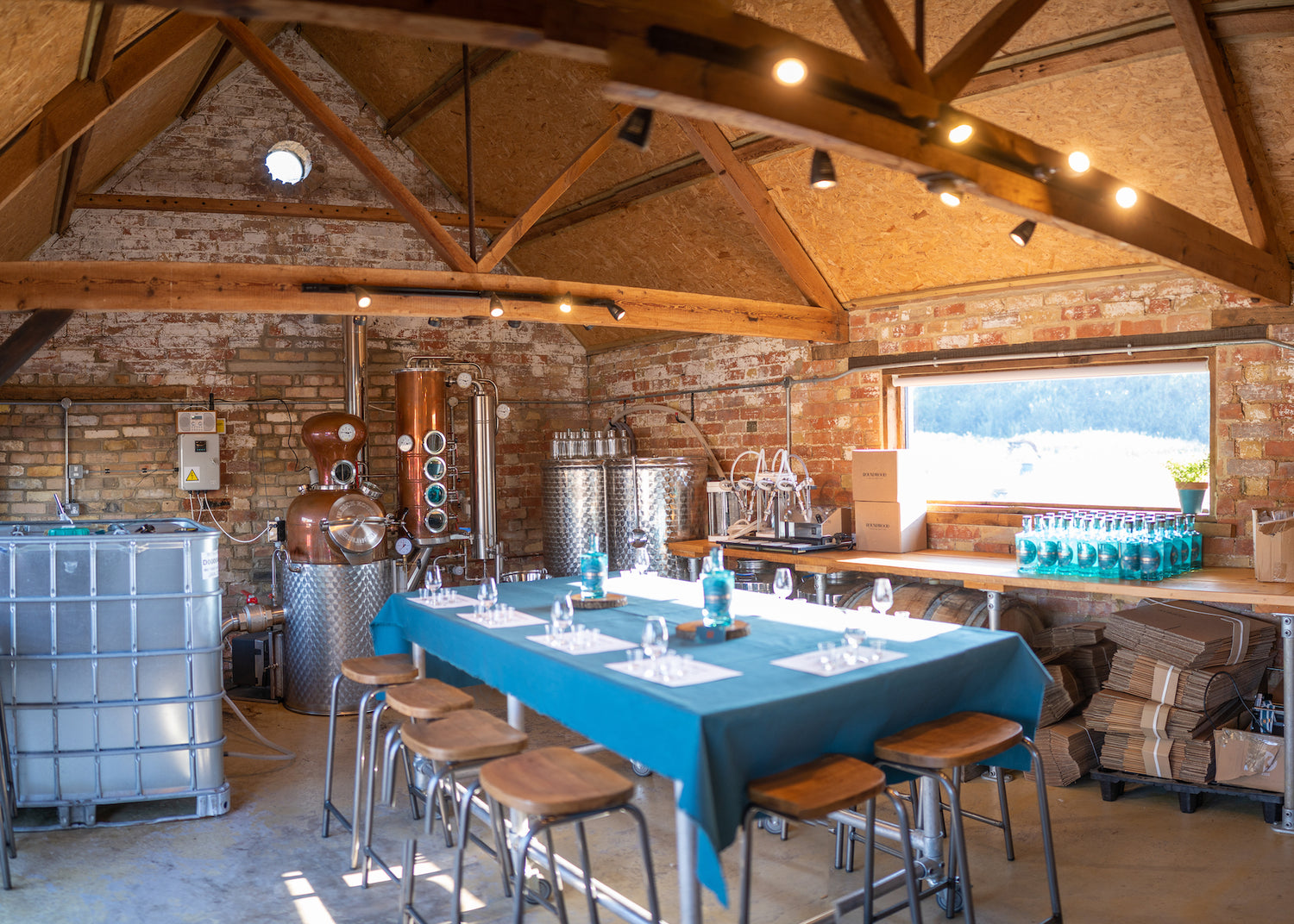 Interior of the small artisan distillery. Old brick barn, with long window and countryside views. Copper still Bluebelle stands at one end and large wooden table in the centre of the distillery, is set for a tasting evening.