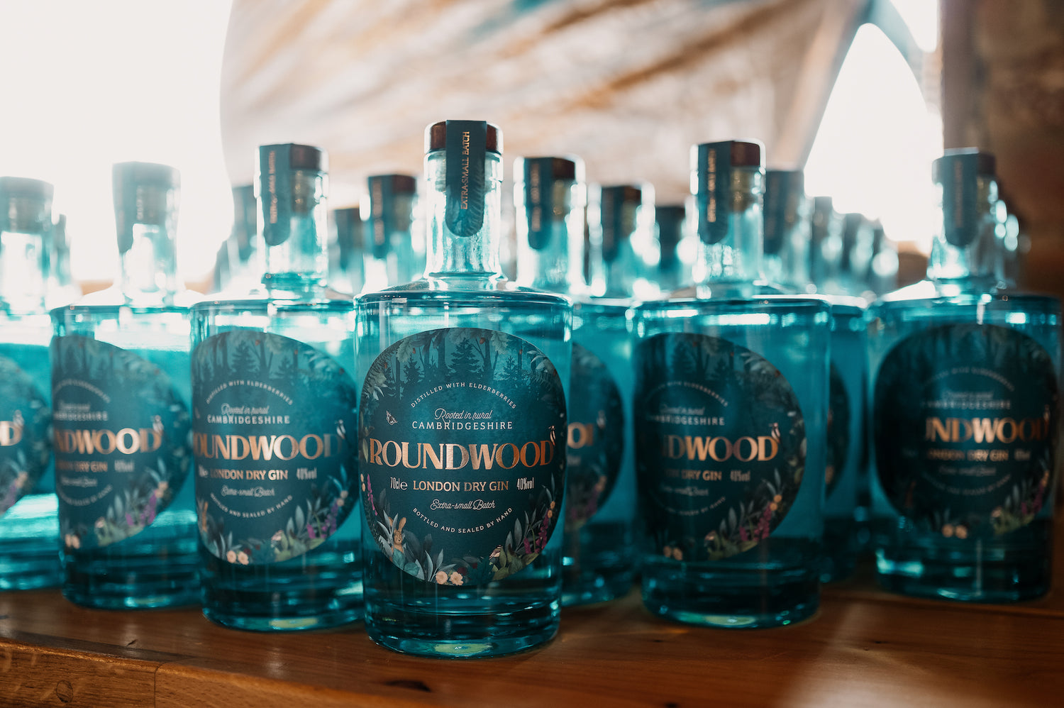 Lots of blue Roundwood Gin bottles stood on wooden bench. Light is shining from behind through distillery window.
